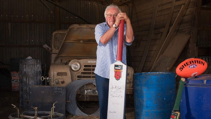 An elderly man stands in a garage leaning on an oversized cricket bat.