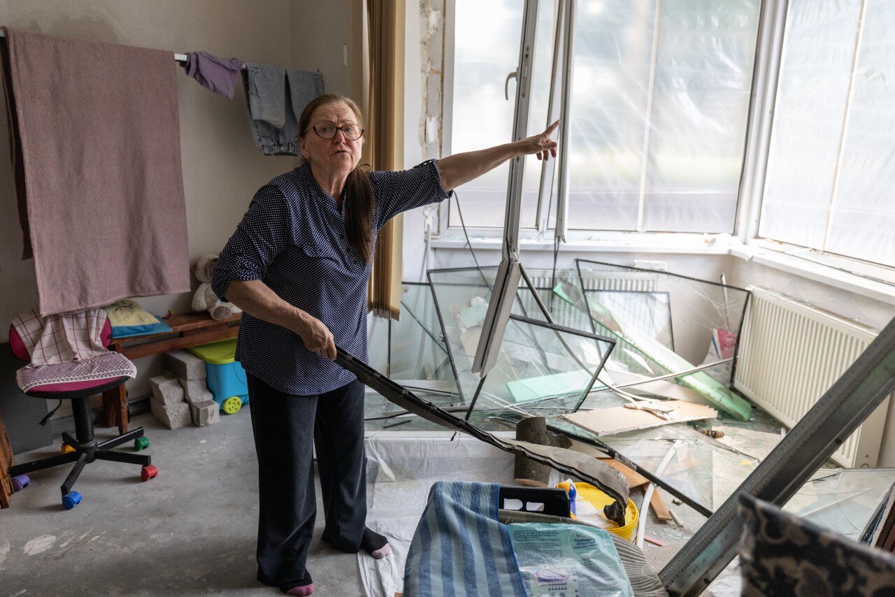 A woman inside the living room of a damaged apartment, pointing at a broken window and looking at the camera
