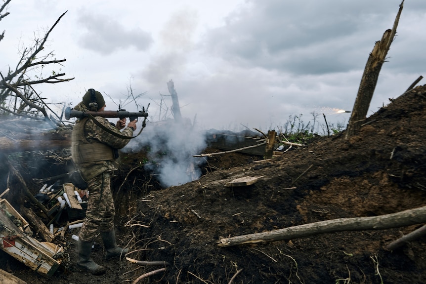 A soldier stands braced with an RPG, a fired rocket flying through the air, a scene of devastation behind him.
