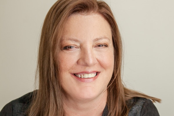 Woman with shoulder-length red hair, broad smile and black jacket. One arm is crossed over her torso, hand resting on shoulder.