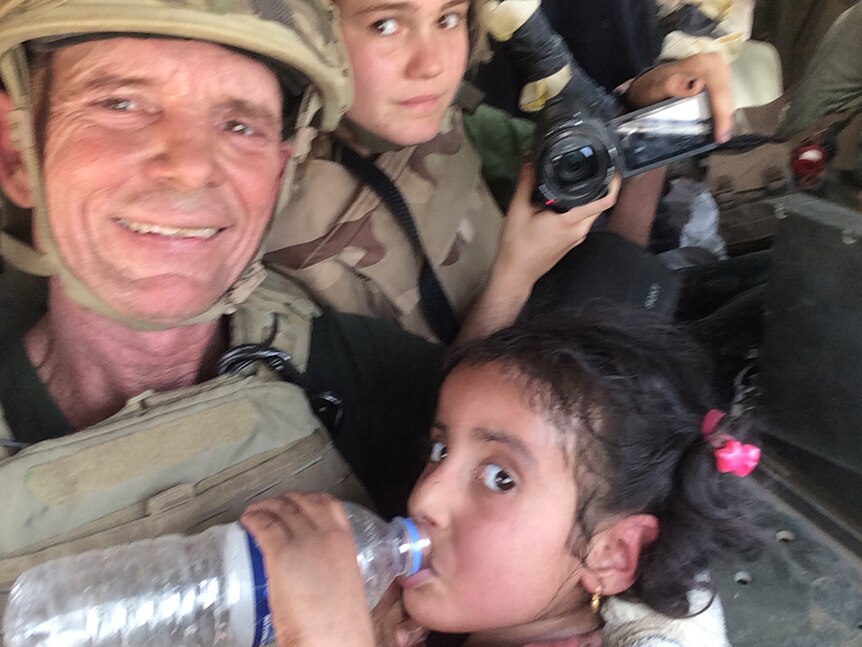 Dave and Sahale Eubank in army fatigues and helmets, with a little Iraqi girl drinking from a water bottle