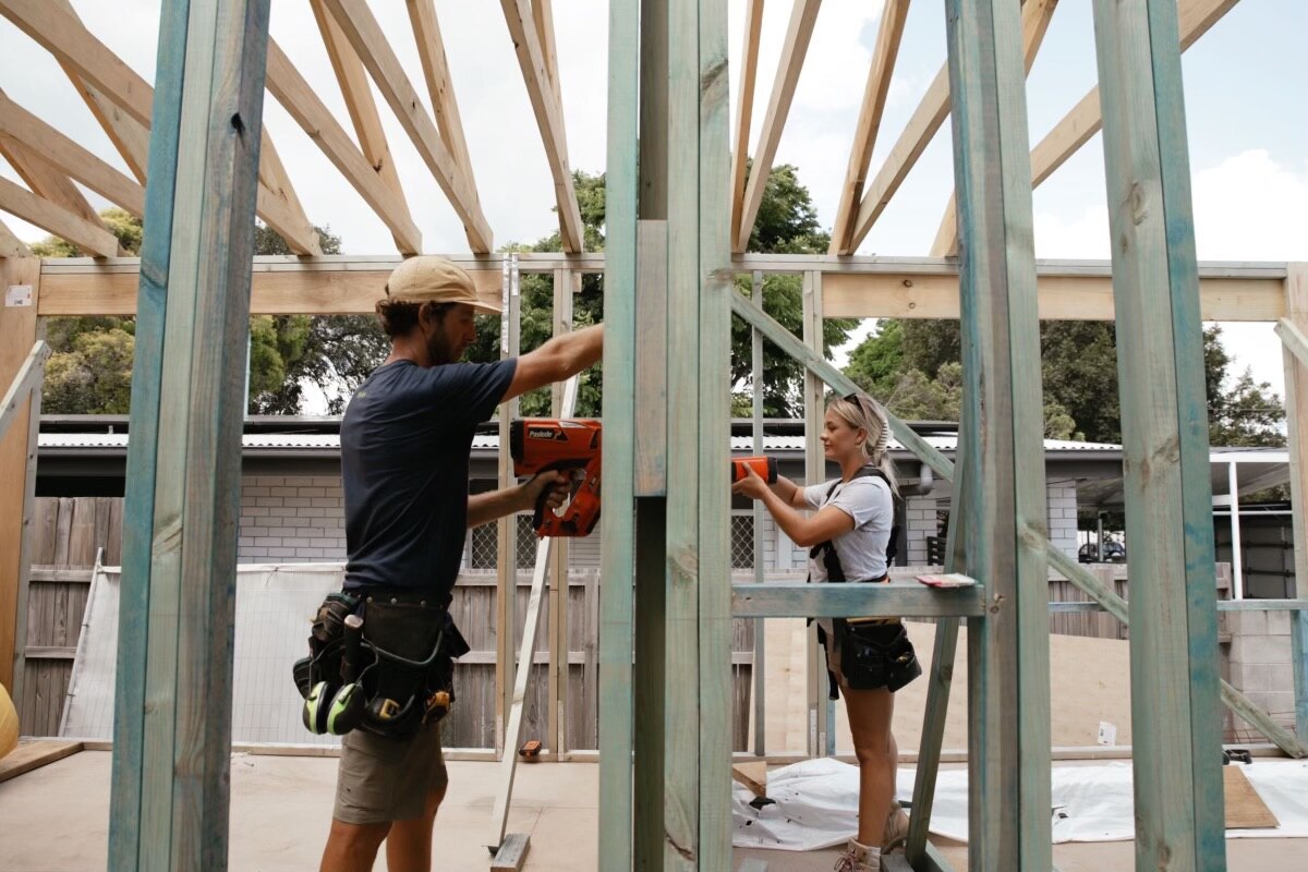 A man wearing a blue T-shirt and a carpentry belt and a woman with a grey T-shirt work on the partly built frame of a house..