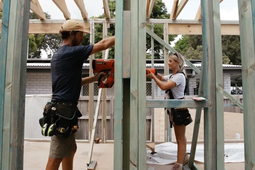 A man wearing a blue T-shirt and a carpentry belt and a woman with a grey T-shirt work on the partly built frame of a house..