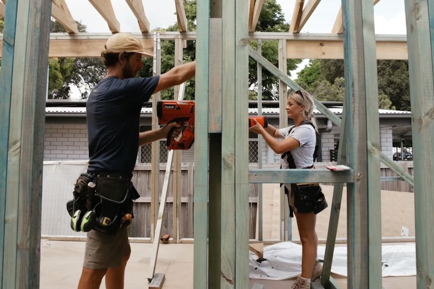 A man wearing a blue T-shirt and a carpentry belt and a woman with a grey T-shirt work on the partly built frame of a house..
