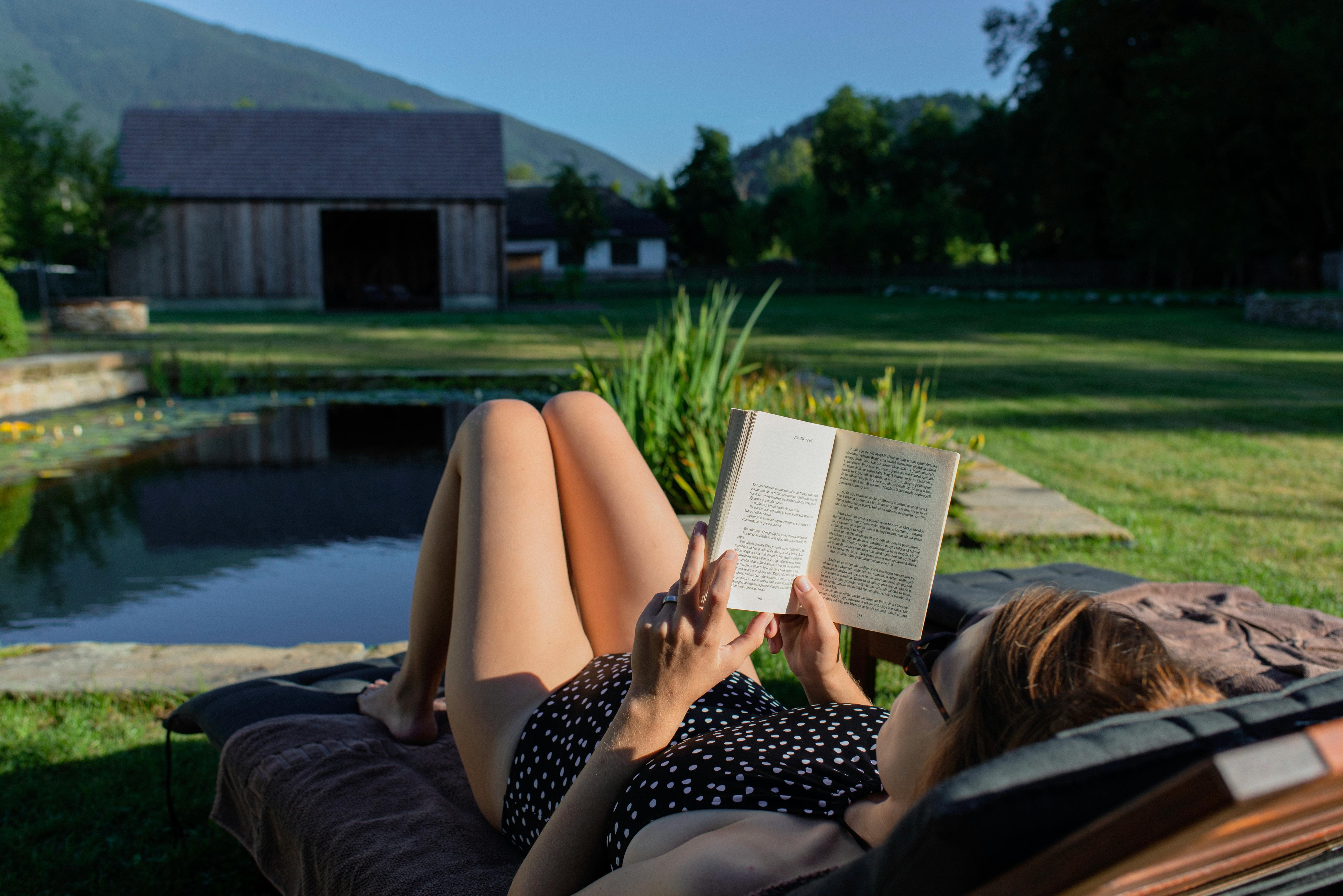 A woman lies by a pond reading a book