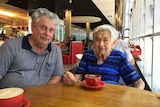Greg Newlyn sitting at a cafe table with his mother Norma, who has a cup of coffee in front of her.