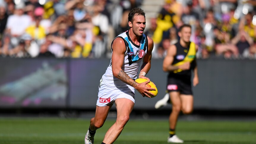 A Port Adelaide AFL footballer holds the ball in both hands as he runs and looks downfield.
