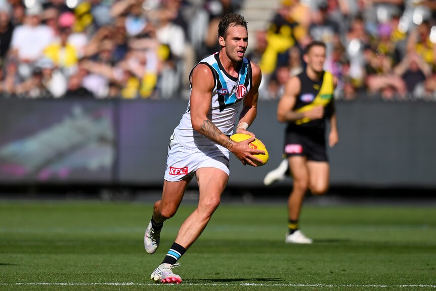 A Port Adelaide AFL footballer holds the ball in both hands as he runs and looks downfield.