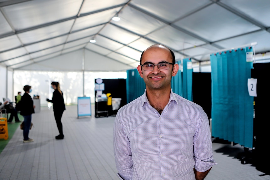 Man with glasses stands smiling inside marquee tent with vaccination booths behind him 