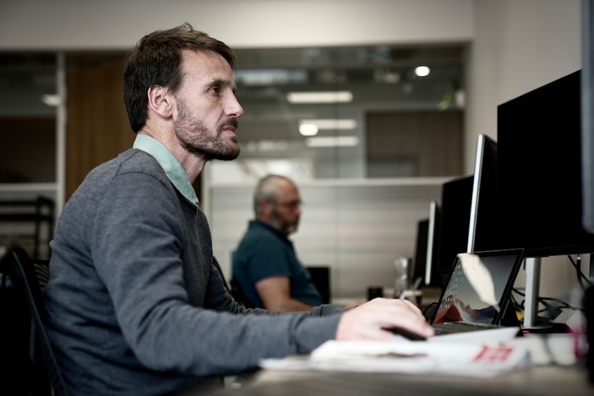 A photo from the side of a man with a beard sitting at a computer. He's wearing a grey jumper.