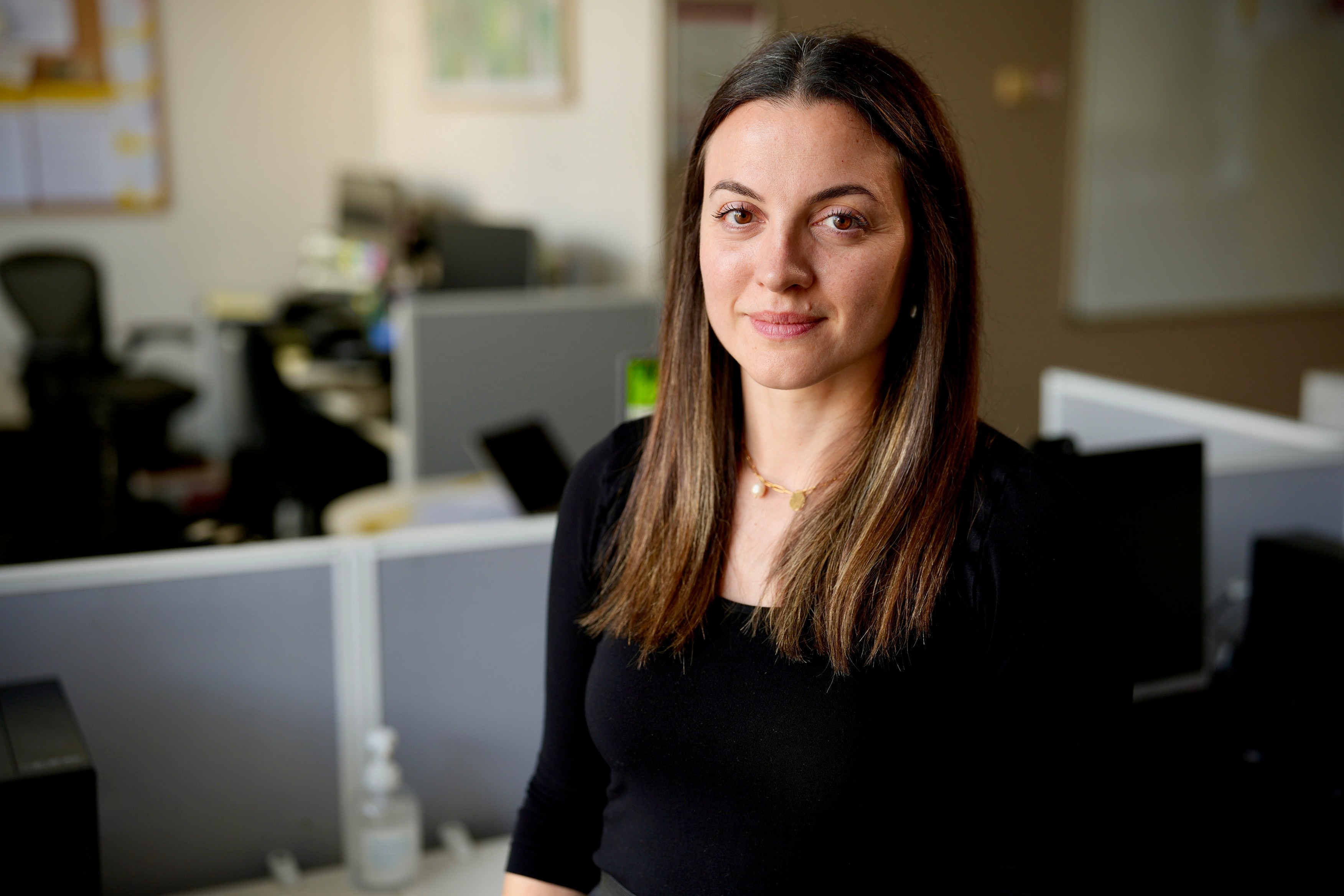 A lady with long brown hair wearing a black top sitting in an office at desks 