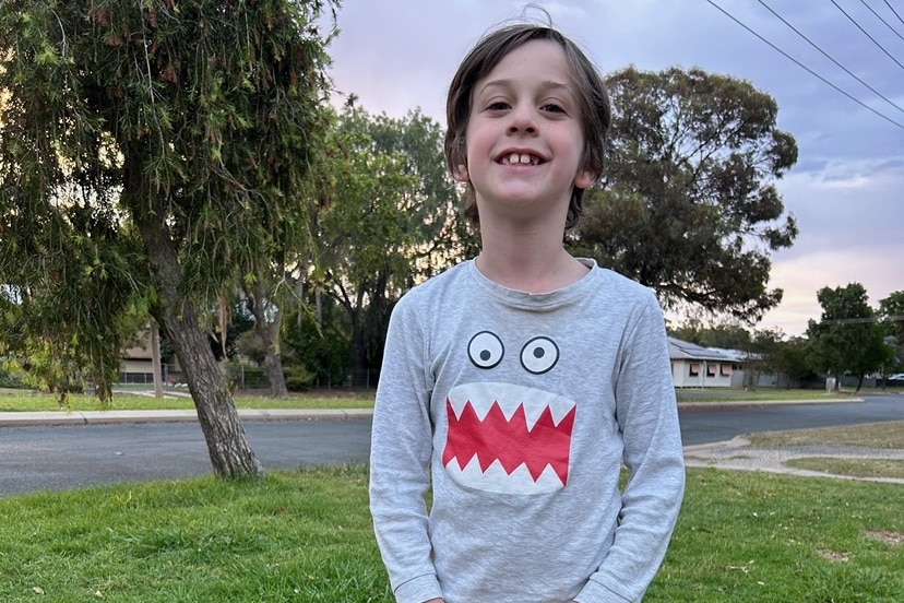 A boy with floppy brown hair smiling wearing a grey long sleeve tshirt with cartoon monster eyes and razor teeth.