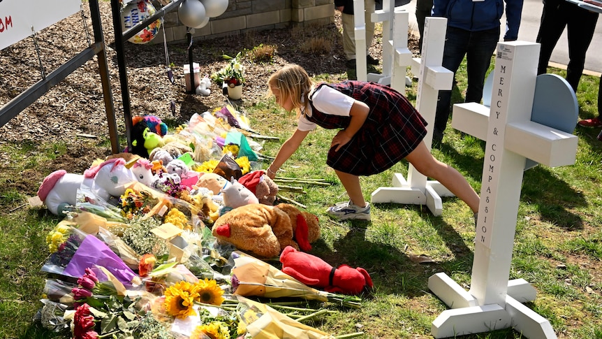 A young girl places a bunch of flowers on a pile of flowers and plush toys behind crosses.