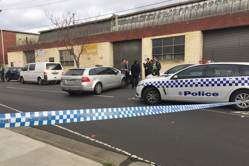 Police gather in a circle while police tape cordons off an area of a warehouse.