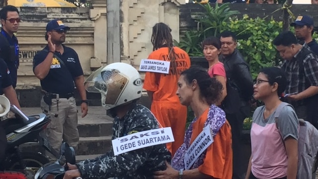 Sara Connor arrives at Kuta Beach on the back of a motorbike surrounded by police and onlookers, David Taylor walks behind her.