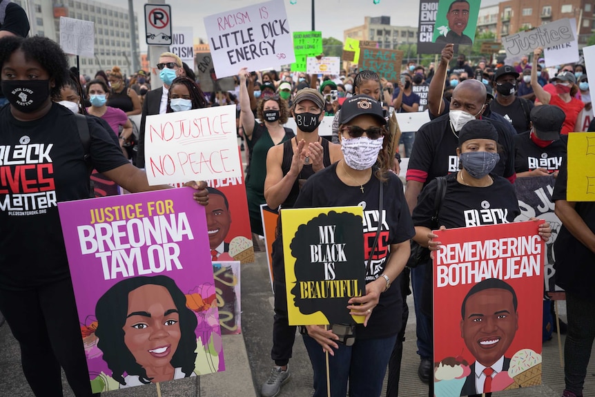 A number of people stand on a street wearing black clothes and masks and holding signs