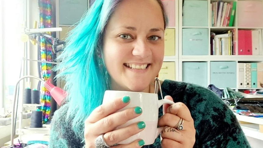 A woman with colourful blue and purple hair smiling holding a mug.