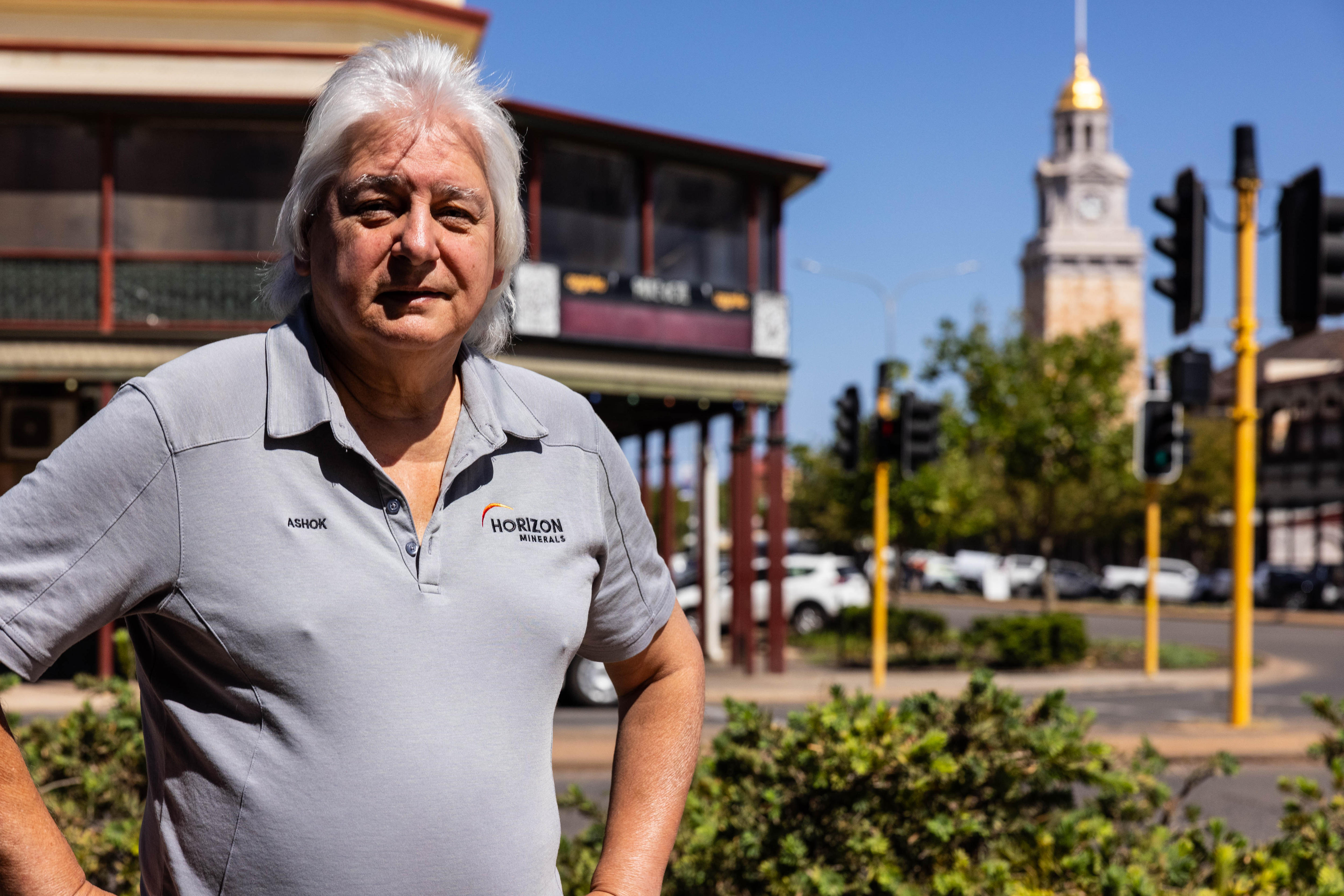 Un hombre mayor, de cabello plateado y con una camiseta polo, se encuentra frente a un pub en un distrito comercial de aspecto histórico.