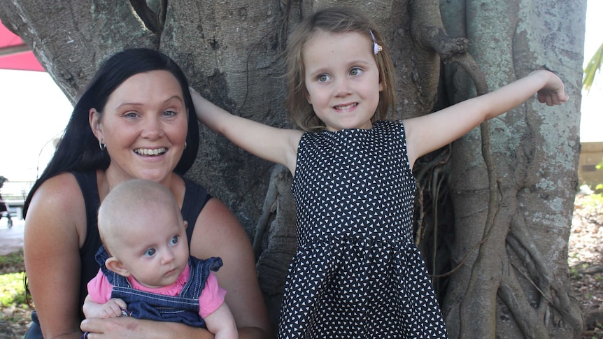 A woman, a baby, and a young girl crouch down under a tree