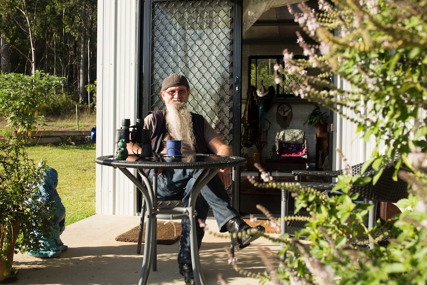 A man with along beard and wearing a cap and glasses sits on a small porch table