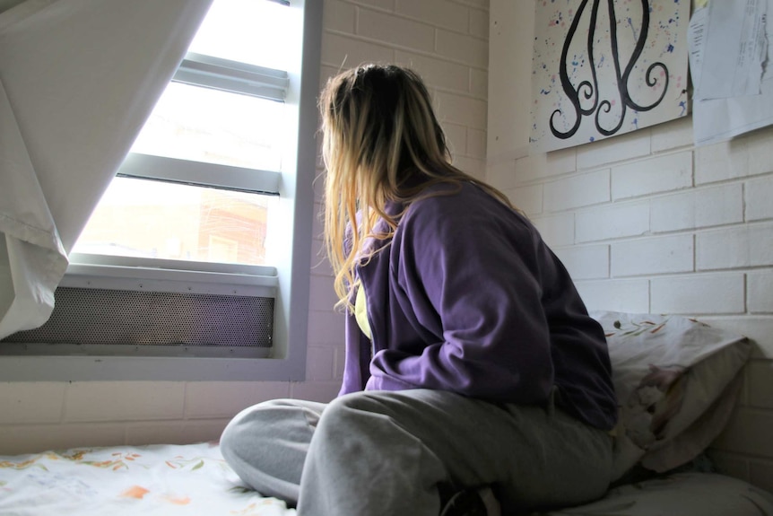 A female prison inmate sits on a bed in a cell wearing a purple jacket and grey pants looking out a window.