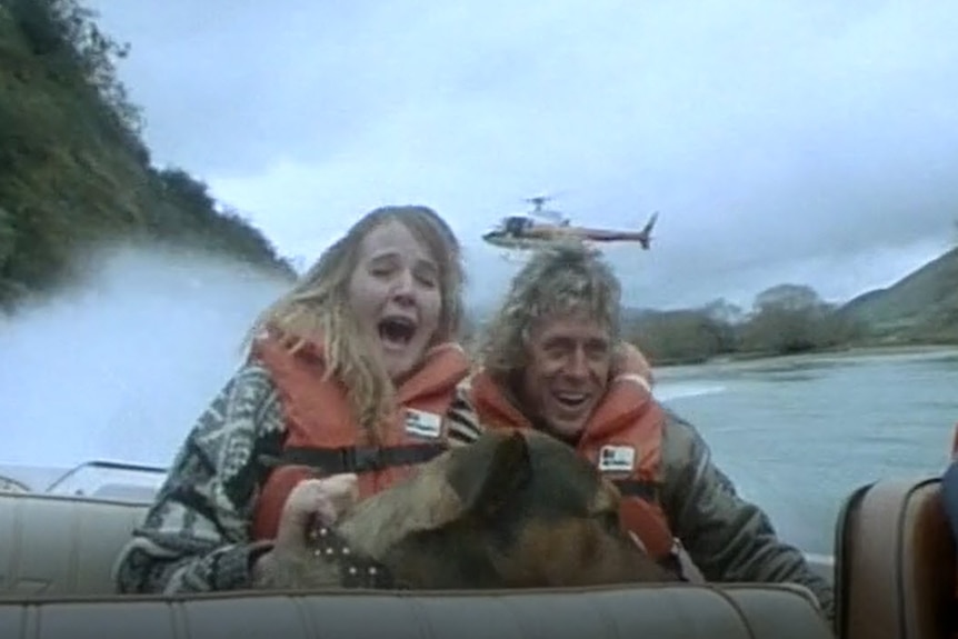 Alby Mangels rides a jet boat with a woman at his side and a helicopter in the background.