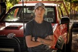 Jai Beaven, wearing baseball cap and black t-shirt, leans on a red 4WD ute with his arms folded across his chest
