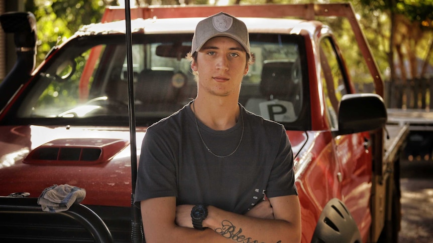 Jai Beaven, wearing baseball cap and black t-shirt, leans on a red 4WD ute with his arms folded across his chest