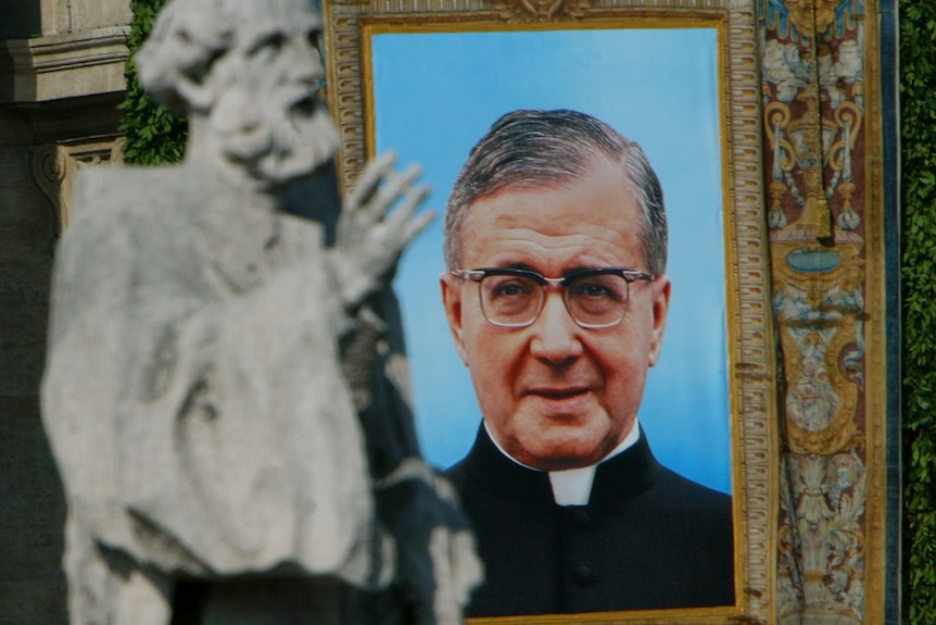 A white man with grey hair in a priest's uniform is shown in a tapestry picture behind a stone statue.