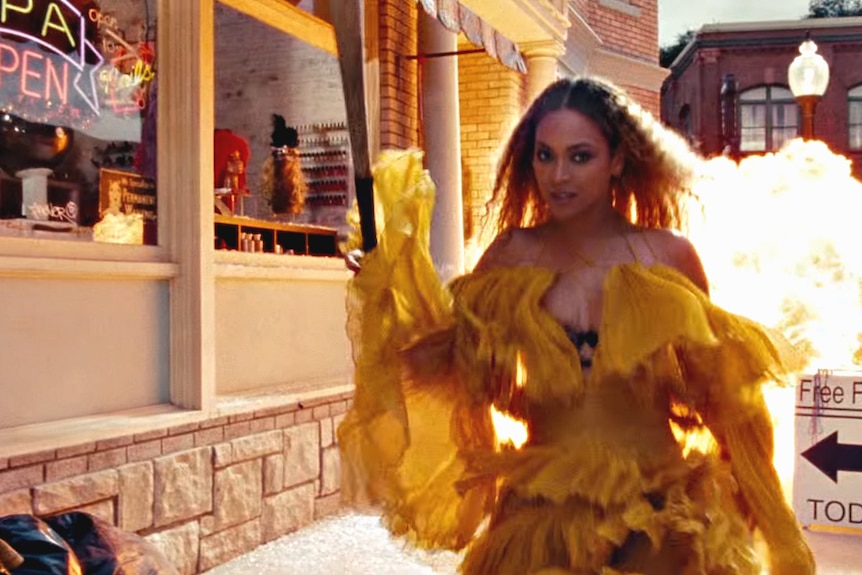 Pop star Beyonce walks down the street in a music video holding a baseball bat in a yellow dress