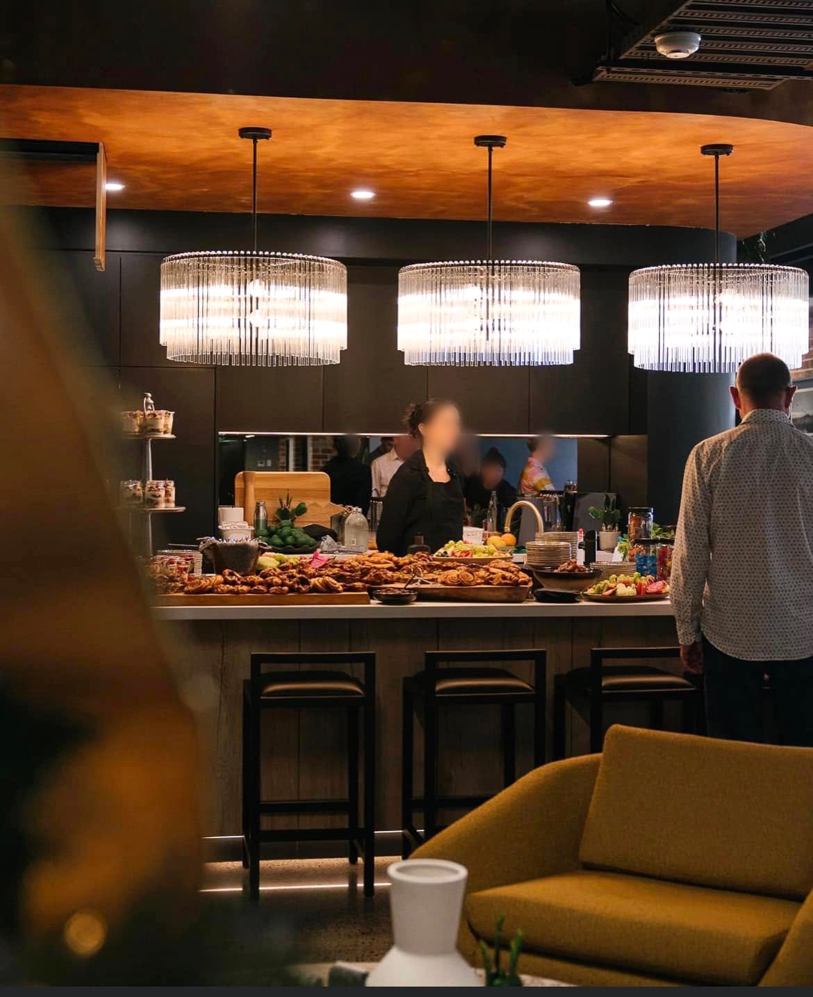 People stand around a large kitchen island covered with plates of food. Above it are three large light fixtures.