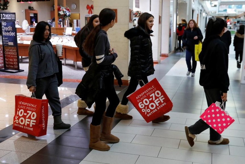 A group of women carrying red shopping bags with SALE signs on them walk through a shopping centre