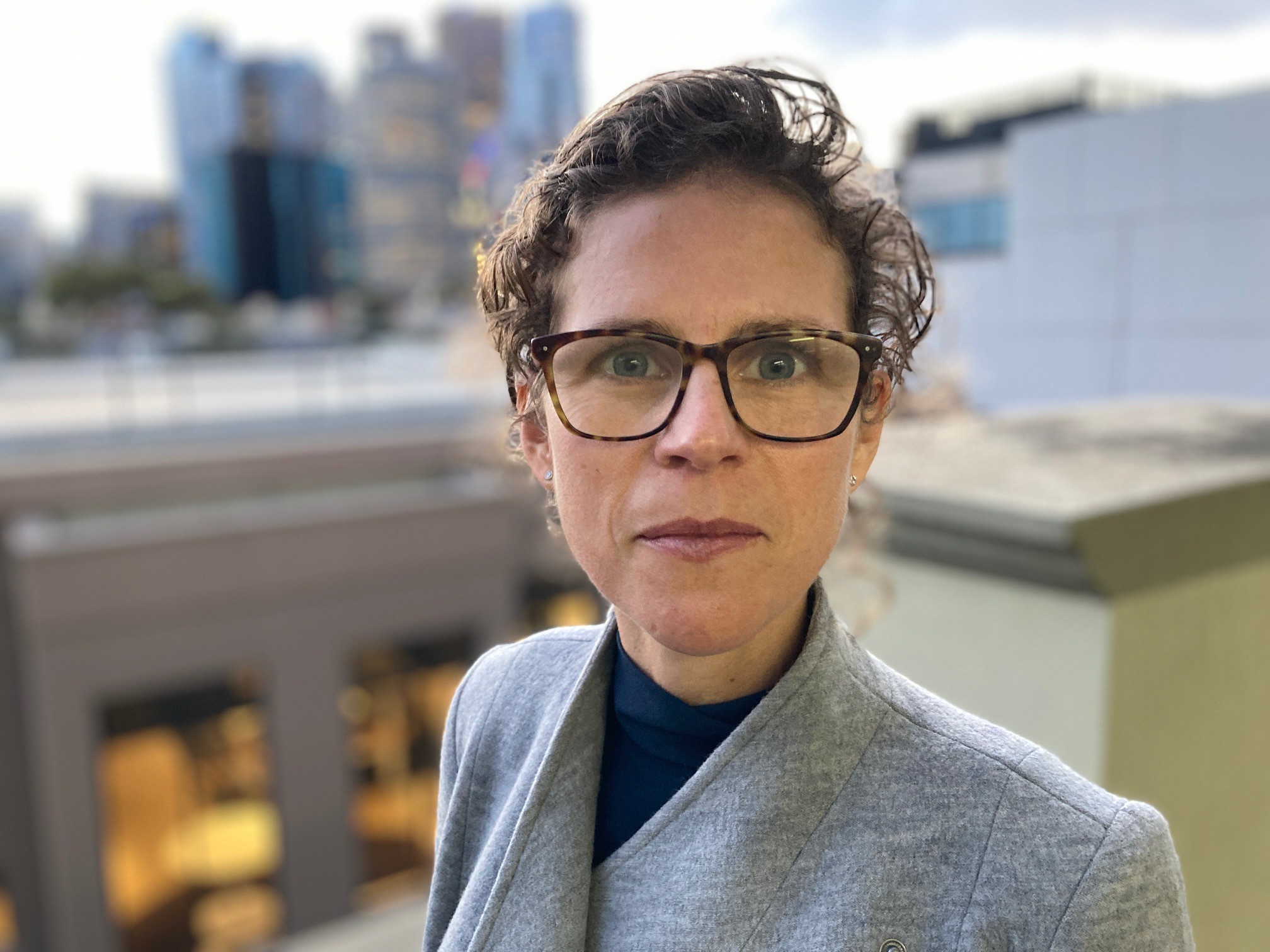 A woman wearing glasses looks straight at the camera with the city in the background.