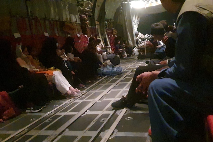 A group of people sit on opposite sides inside a cargo plane with light streaming in from distance.