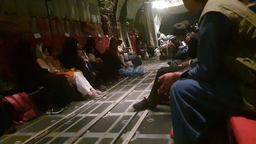 A group of people sit on opposite sides inside a cargo plane with light streaming in from distance.
