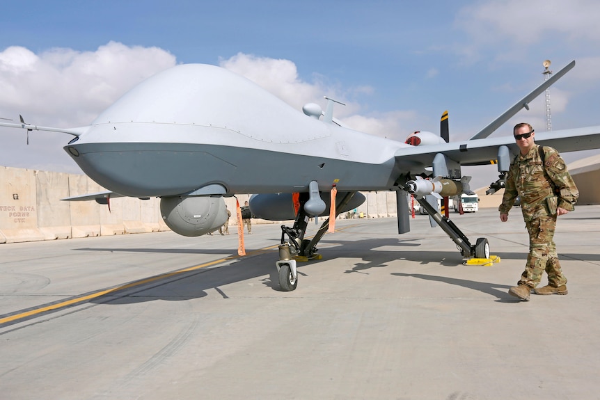 A man in camo gear walks next to a big grey aircraft