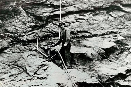 A black and white photo of a man standing on a rocky ledge beside a long piece of rope.