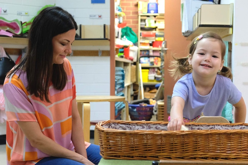 Maya Linden kneels by her daughter Lily, who is pulling a toy out of a basket.