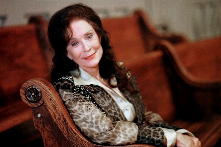 Country music singer Loretta Lynn poses for a portrait, sitting in a large chair and smiling.