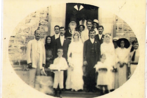 An yellow faded photograph of two sisters in white wedding dresses and their grooms, surrounded by family members.