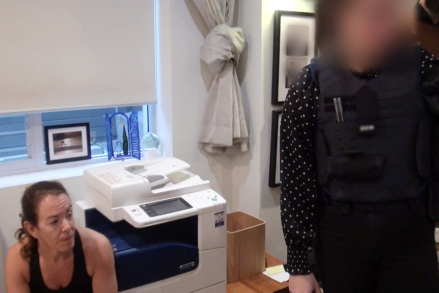 a woman sitting down next to a photocopier looking as police examine her belongings