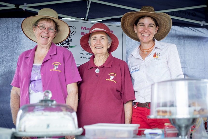 Three women wearing wide-brimmed hats stand behind a display of cakes at a country market stall.