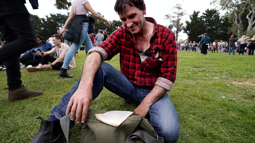 A concert-goer sits on grass reading a book.