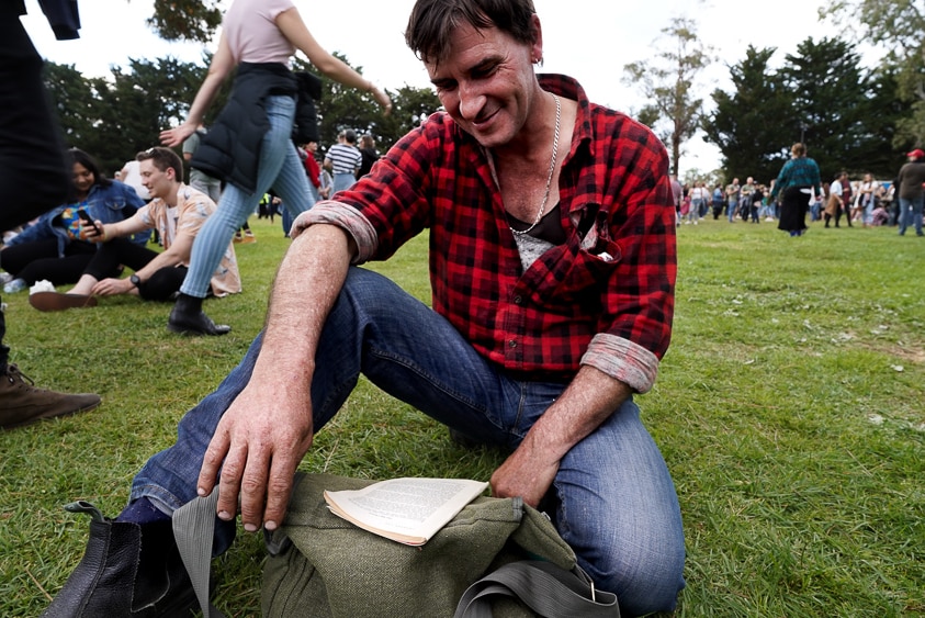 A concert-goer sits on grass reading a book.