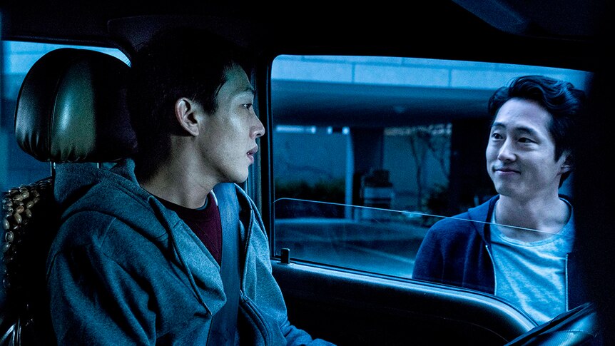 Colour still of Yoo Ah-in sitting in car and Steven Yeun standing outside car window in 2018 film Burning.