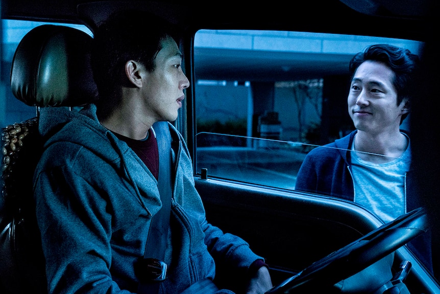 Colour still of Yoo Ah-in sitting in car and Steven Yeun standing outside car window in 2018 film Burning.