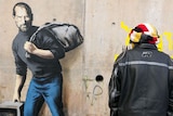 Man stands in front of mural of man holding an old computer and bag over his shoulder