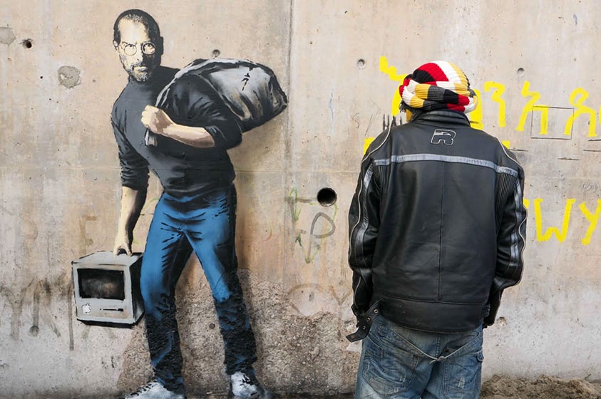 Man stands in front of mural of man holding an old computer and bag over his shoulder