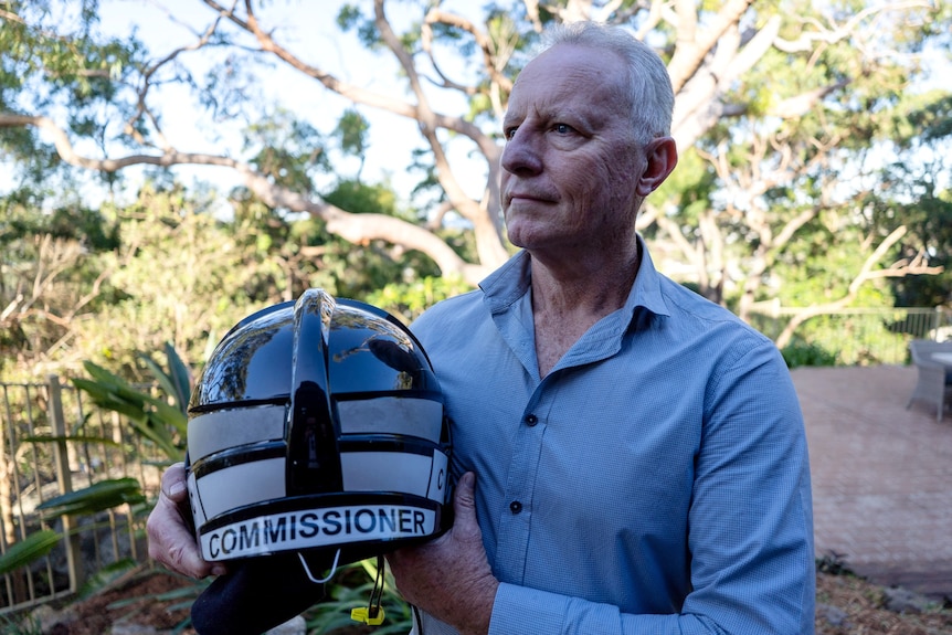 Greg Mullins holds a helmet stamped with the word "Commisioner", looking into the distance in front of a leafy area.