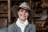 A smiling woman in a felt hat and jumper in a shed. 
